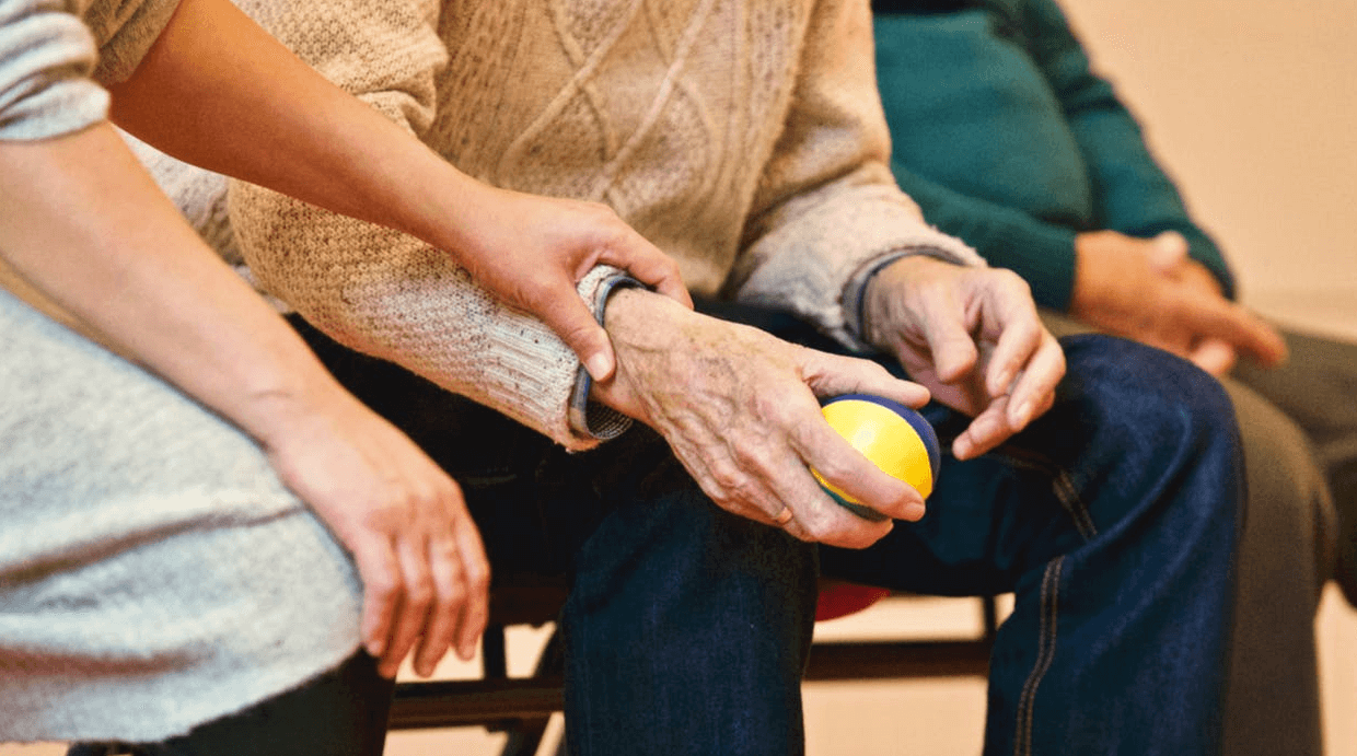 A younger person holding a senior citizen's hand as he grips a ball
